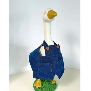 May include: A white goose ornament with a yellow beak and feet, wearing a blue denim overall. The overall has two pockets and brown buttons. The goose stands on a green base.