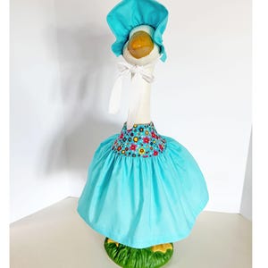 May include: A decorative goose figurine wearing a turquoise bonnet and dress. The dress features a floral pattern around the neckline. The goose stands on a green base, creating a whimsical and charming aesthetic.