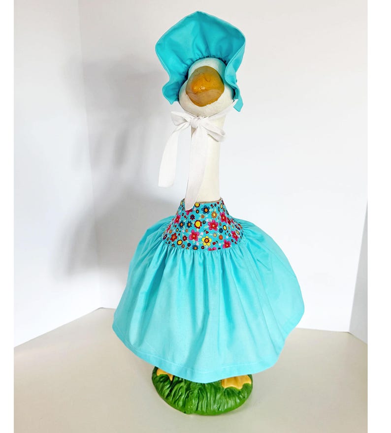 May include: A decorative goose figurine wearing a turquoise bonnet and dress. The dress features a floral pattern around the neckline. The goose stands on a green base, creating a whimsical and charming aesthetic.