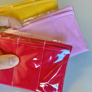 May include: Three fabric items in red, pink, and yellow, each encased in clear plastic. The items have a rectangular shape with vertical stitching. The red item is in the foreground, with the pink and yellow items behind it.