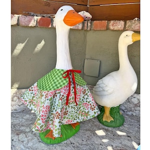 May include: Two white goose statues with orange beaks and feet standing on green bases. The larger goose wears a green and white checkered capelet over a white skirt featuring a red and green holly berry pattern, tied with a red ribbon.
