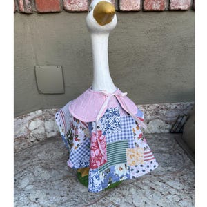 Op de afbeelding: Een wit ganzenbeeld met een goudkleurige snavel draagt een patchwork cape. De cape heeft een lichtroze kraag met witte sterren en een striksluiting, terwijl het lijfje bestaat uit diverse stoffen vierkantjes met bloemen-, ruit- en streepmotieven.