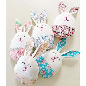 Peut inclure: Plusieurs lapins de Pâques en peluche faits à la main. Chaque lapin a un corps blanc, des traits de visage roses brodés et des accents en tissu à motifs sur les oreilles et le bas du corps. Les motifs de tissu comprennent des motifs floraux.