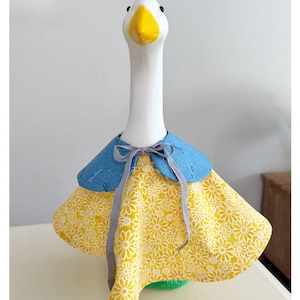 Könnte beinhalten: Eine weiße Gänsefigur aus Keramik, die einen blauen Stoffkragen mit kleinem Scherenmuster und einen gelben Stoffrock mit weißem Gänseblümchenmuster trägt. Das Outfit ist am Hals mit einem dünnen grauen Band zu einer einfachen Schleife gebunden.