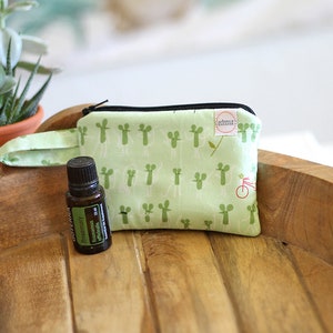 May include: A small green zippered pouch with a white cactus print and a black zipper. The pouch has a green fabric handle and is sitting on a wooden surface next to a small brown bottle labeled "Rosemary Essential Oil".
