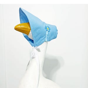 May include: A light blue bonnet with a floral applique, tied with white ribbons. The bonnet is worn by a goose statue with a yellow beak. The bonnet is a whimsical accessory.