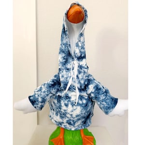 May include: A whimsical goose figurine wearing a blue and white tie-dye hoodie with white drawstrings. The goose has an orange beak and feet, and white wings. The base of the goose is green.
