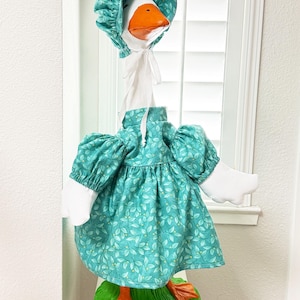 Könnte beinhalten: Dekorative Gänse-Statue mit teal-farbenem Kleid und Haube mit Blättermuster. Die Gans hat einen orangefarbenen Schnabel und Füße und steht auf einem grünen Sockel. Ein skurriles Wohnaccessoire.