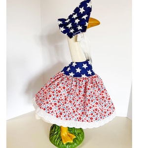 May include: A goose statue wearing a patriotic outfit. The outfit includes a blue bonnet with white stars, a matching collar, and a red floral skirt with a white lace trim. The goose stands on a green base.