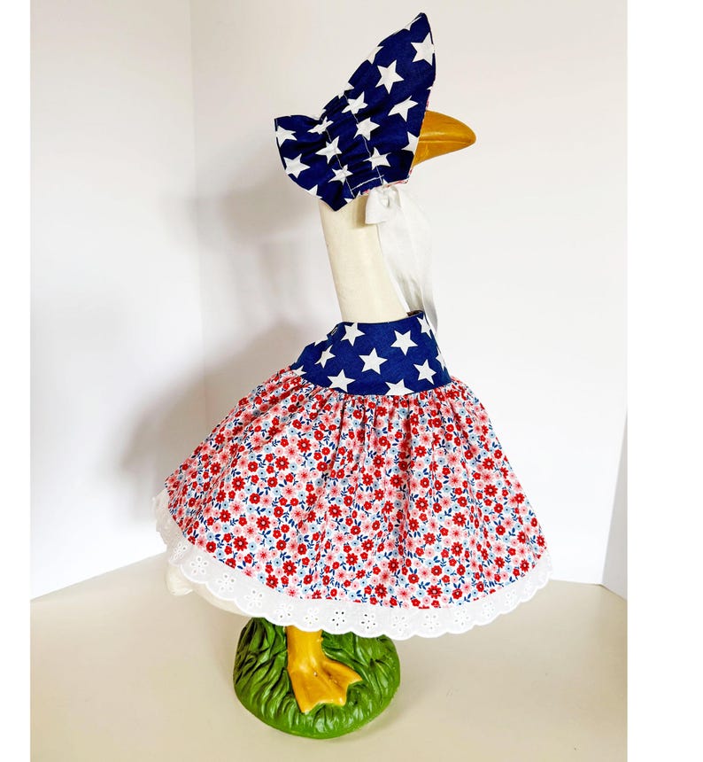 May include: A goose statue wearing a patriotic outfit. The outfit includes a blue bonnet with white stars, a matching collar, and a red floral skirt with a white lace trim. The goose stands on a green base.