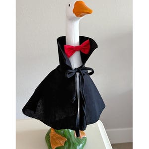 Può includere: Una statuetta di oca bianca con becco e zampe arancioni su una base verde. L'oca indossa un mantello nero con colletto alto e chiusura a laccio nero sul collo, impreziosito da un papillon rosso brillante attorno al collo bianco.