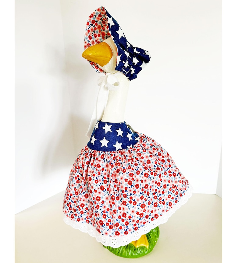 May include: A decorative goose figurine wearing a patriotic outfit. The outfit includes a blue and white star-patterned bonnet and a red floral print skirt with a blue star-patterned waistband. The skirt has a white lace trim.