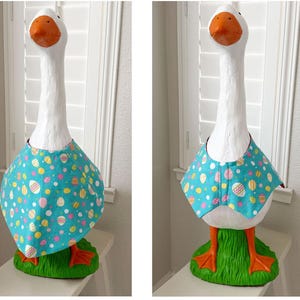 May include: A white goose statue with an orange beak and feet, wearing a turquoise bib adorned with colorful Easter egg patterns. The goose stands on a green grassy base, perfect for Easter decor.