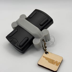 May include: A black and gray car-shaped phone holder with a suction cup on the back. The holder has a small wooden tag attached with the letter "W" and a wing design.