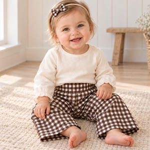 May include: A long-sleeved cream-colored shirt paired with brown and white gingham pants. The outfit includes a matching brown and white gingham bow headband. The shirt features a simple round neckline and long sleeves with gathered cuffs.