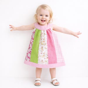 May include: A patchwork dress with a pink neckline and hem. The dress features panels of floral, polka dot, bunny print, pink, and gingham fabrics. The child is wearing white sandals.
