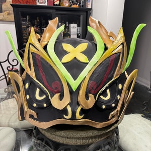 Könnte beinhalten: Eine schwarze, goldene und grüne Drachenmaske mit aufwendigen Details. Die Maske hat ein großes, gelbes Blumenmuster auf der Stirn und ein Paar große, gelbe Augen.