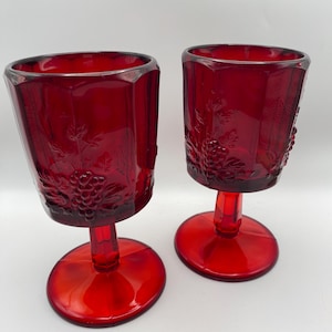 May include: Two ruby red vintage goblets with a textured grape and leaf design. The goblets have a hexagonal shape and sit on a round base. The stem is short and the glass is a deep, rich red colour.