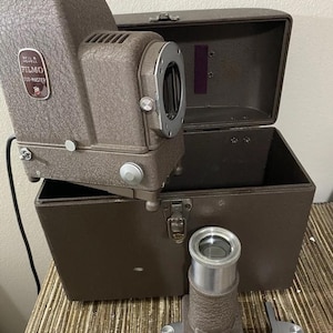 May include: A vintage brown Bell & Howell Filmo 126-Master slide projector in a brown carrying case with a lens attached.