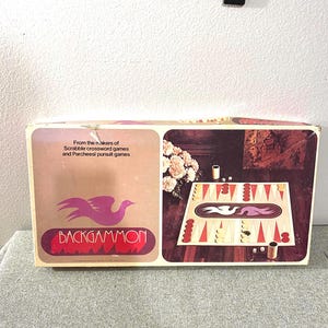 Puede incluir: Una caja de juego de backgammon vintage con fondo marrón y un diseño de pájaro blanco. La caja presenta el texto "From the makers of Scrabble crossword games and Parcheesi pursuit games" y "Backgammon".