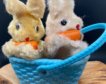 May include: Two plush bunny rabbits holding orange carrots sit inside a blue plastic basket with a woven design. The basket has a blue handle.