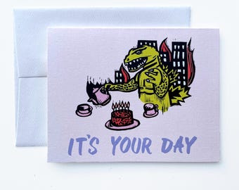 May include: A greeting card with a light purple background and a matching envelope. The card features a green monster pouring tea, with a cake and cups. The text "IT'S YOUR DAY" is printed in blue.