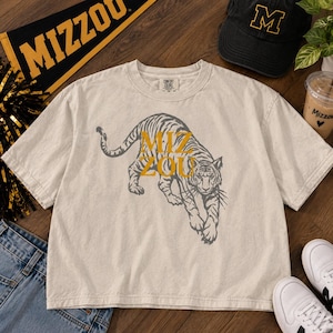 May include: A beige short-sleeve t-shirt featuring a dark grey graphic of a tiger in a prowling stance. The text MIZ ZOU is printed in gold letters over the tiger's body. The shirt has a crew neckline and a label at the inner neck area.
