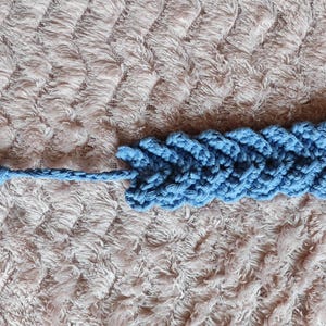 May include: A blue crocheted bookmark with a tassel. The bookmark is made of a soft, textured yarn and has a simple, elegant design.