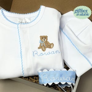 May include: White baby outfit set with light blue trim, including a onesie, hat, and bib. The onesie features an embroidered teddy bear and the name "Rowan" in light blue script. The set is presented in a gift box.