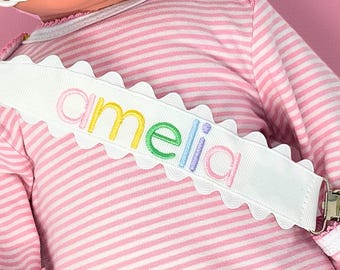 Puede incluir: Pinza para chupete blanca con el nombre "Amelia" bordado en colores arcoíris. El clip tiene un borde festoneado y un cierre metálico. El fondo es una camisa a rayas rosas y blancas.