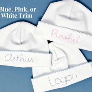 May include: Three white baby hats with folded brims and scalloped edges. The hats feature embroidered names in blue or pink thread. The text "Blue, Pink, or White Trim" is visible at the top. The hats are made of a smooth, solid white fabric.