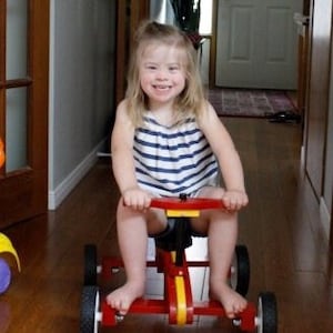 May include: A young girl with blonde hair is smiling while riding a red and yellow toy car with black wheels. The car has a yellow stripe down the middle and a black seat.