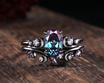 May include: A black gold ring set with a large oval-shaped blue and purple gemstone surrounded by smaller clear and purple gemstones. The ring has a delicate vine design.