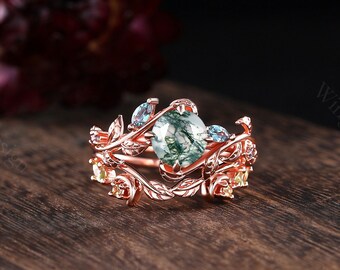 May include: A rose gold ring set featuring a moss agate center stone, accented with blue and yellow gemstones. The ring has a floral design with intricate leaf and vine details. The set is displayed on a wooden surface.