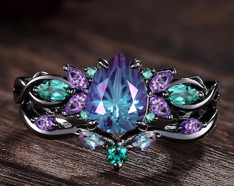 May include: A black ring with a large, pear-shaped, teal and purple gemstone at the center, surrounded by smaller teal and purple stones. The ring is set on a wooden surface, showcasing the intricate design and color contrast.