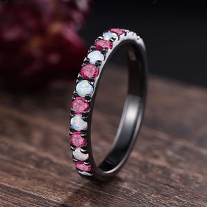 May include: A black ring with a row of alternating pink and white gemstones. The ring is set on a wooden surface, with a dark background. The gemstones are small and evenly spaced around the band.