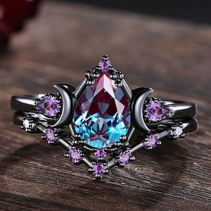 May include: A black ring set featuring a pear-shaped, color-changing gemstone with purple and teal hues. The ring is accented with crescent moon and small purple gemstones. The design is set on a dark wooden surface.