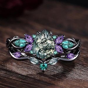 May include: A black ring with a central oval-shaped moss agate stone, surrounded by teal and purple gemstones. The ring has a detailed, ornate design, showcasing a nature-inspired aesthetic. The ring is set on a wooden surface.