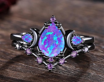 May include: A black ring set featuring a pear-shaped, iridescent blue-purple opal center stone. The ring is accented with small purple gemstones and crescent moon details. The set is displayed on a wooden surface.