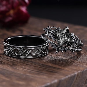 May include: Two black rings on a wooden surface. One ring has a floral design, the other has a large, faceted stone and smaller black stones. The rings are likely for a wedding or engagement.