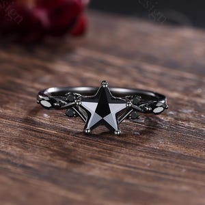 May include: A black star-shaped ring with a faceted black gemstone center. The ring has a dark metal band with leaf and small black stone accents. The ring is set on a wooden surface.