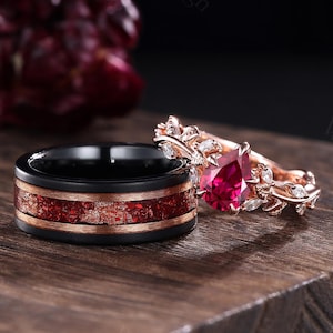 Peut inclure: Deux bagues sur une surface en bois. L'une est une alliance noire avec une incrustation rouge et dorée. L'autre est une bague en or rose avec une pierre rouge en forme de poire et des accents de feuilles. L'arrière-plan est sombre.