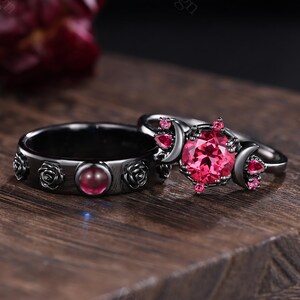 May include: Two black rings with red gemstones. One ring features a central red gemstone surrounded by rose designs. The other ring has a large, round red gemstone and smaller red gems.