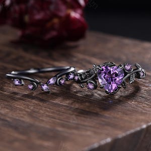 May include: A black engagement ring set featuring a heart-shaped purple gemstone and smaller round gemstones. The rings have a leaf and vine design. The set is displayed on a wooden surface.