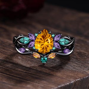 May include: A black ring with a large, pear-shaped yellow gemstone at the center, surrounded by smaller teal, purple, and orange gemstones. The ring's band has a detailed, ornate design. The ring is displayed on a dark wooden surface.