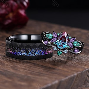 May include: Two rings on a wooden surface. One ring is black with a colorful, speckled design. The other ring is black with a large, pear-shaped, multi-colored gemstone and smaller surrounding gemstones.