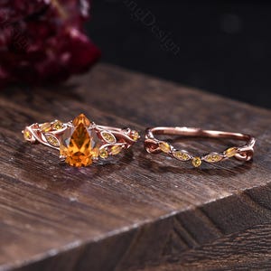 May include: A rose gold ring set featuring a pear-shaped citrine center stone flanked by smaller yellow stones set in a leaf-inspired band, accompanied by a matching curved band with small yellow stones, both crafted in a polished metal finish.