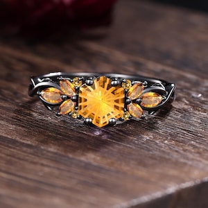 May include: A black ring with a large, hexagonal, yellow gemstone at the center, flanked by smaller, leaf-shaped orange gemstones. The ring is set on a dark wooden surface, showcasing the jewelry's design and color contrast.
