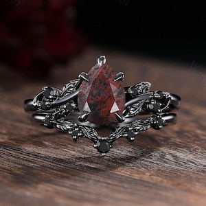 May include: A black engagement ring set with a pear-shaped, dark red gemstone. The ring features intricate leaf and black gemstone accents. The band is black and has a matching wedding band.