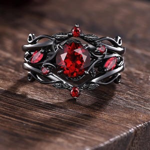 Peut inclure: Une bague en métal noir ornée d'une grosse pierre rouge ronde au centre, entourée de petites pierres rouges et de motifs complexes en forme de feuilles. La bague est posée sur une surface en bois.